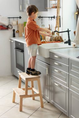 Küçük çocuk mutfakta tabak yıkıyor.