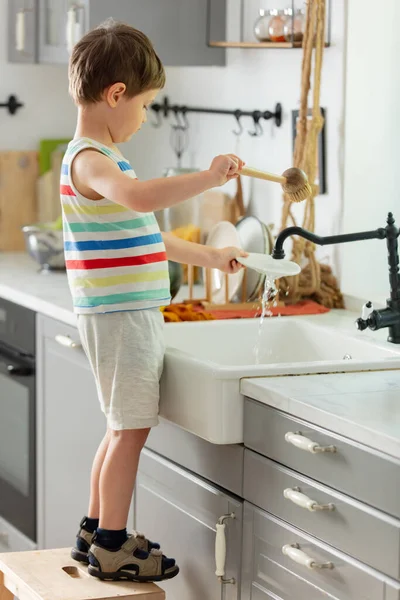 Küçük çocuk mutfakta tabak yıkıyor.