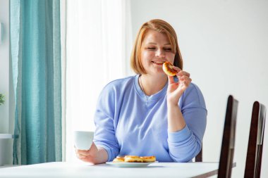 Ukraynalı göçmen kadın kahvaltıda elinde fincan ve peynirli kekle masada oturuyor.