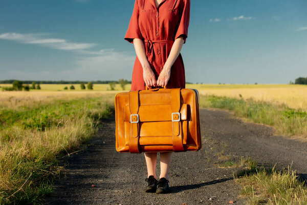 Girl in red dress with suitcase on country road in sunset. Low side view