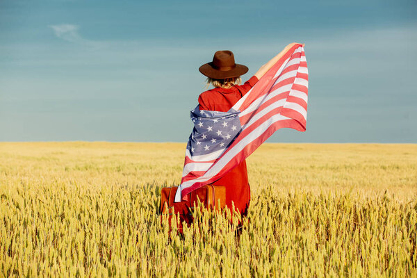 Girl in USA flag with suitcase in wheat field with blue sky on background