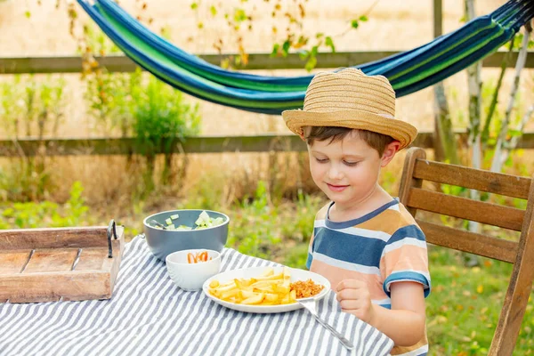 小男孩坐在花园的桌旁 吃着沙拉和薯条 — 图库照片