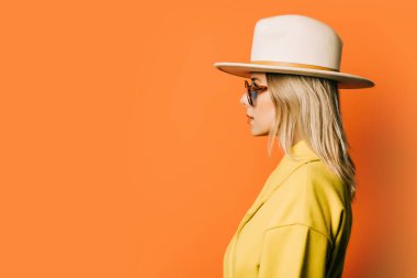 Stylish 80s woman in eyeglasses and hat with jacket on orange background