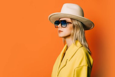 Stylish 80s woman in eyeglasses and hat with jacket on orange background