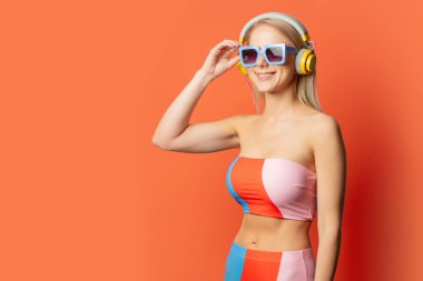Renkli mayo giymiş, kulaklık takan ve turuncu arka planda güneş gözlüğü takan şık sarışın kadın.