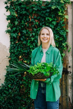 Sepet dolusu sebzeli yeşil gömlekli şık bir kadın sarmaşık arka planında duruyor.