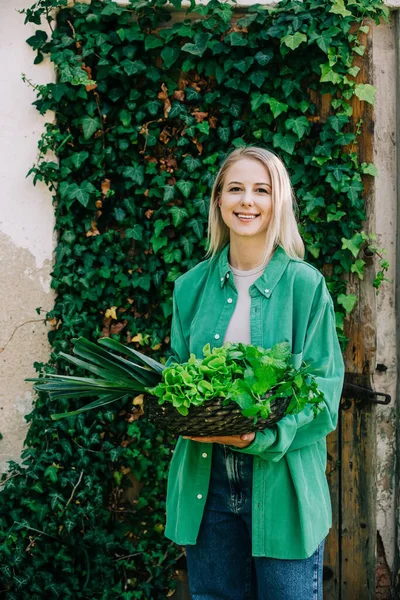 Sepet dolusu sebzeli yeşil gömlekli şık bir kadın sarmaşık arka planında duruyor.