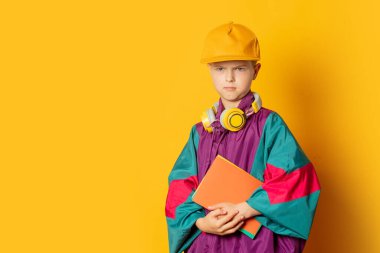 Stylish kid boy in 80s sport suit and cap with notebooks on yellow background