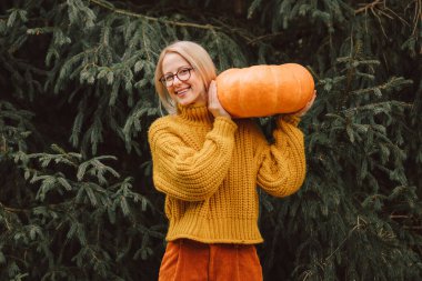 Stylish woman in yellow sweater with pumpkin stands near spruce tree