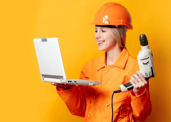 スタイリッシュなブロンドの髪労働者女性でオレンジヘルメットとスーツでドリルとラップトップで黄色の背景 — ストック写真