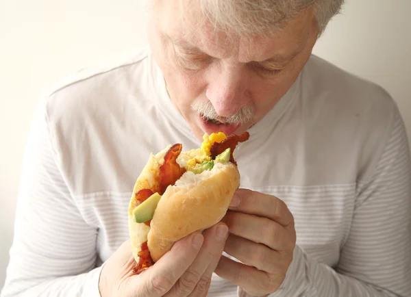 Homem mais velho come bacon, ovo e sanduíche de abacate — Fotografia de Stock