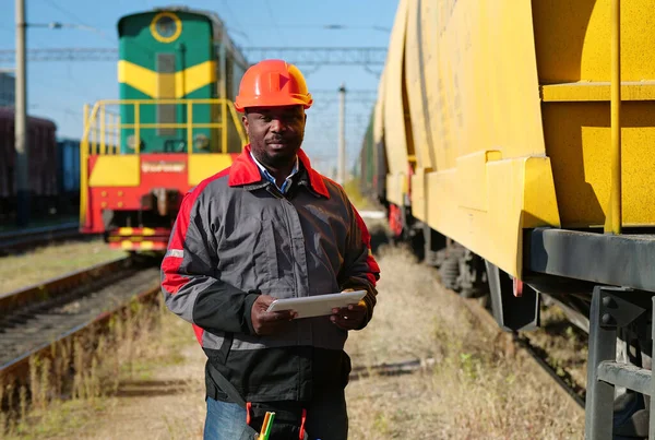 Afro-american railway man with tablet computer at freight train terminal. Railroad man in uniform and red hard hat with computer. Railway employee holds in hands tablet pc and look at the camera