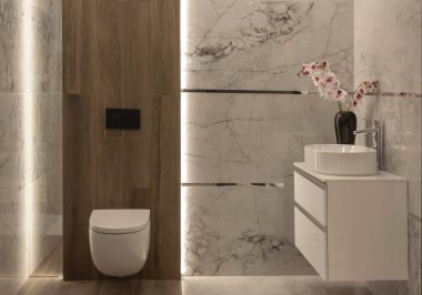 Bathroom with a view of a closed toilet, tap, cupboard, washbasin, flowers in a vase and wood-like tiles on the wall.