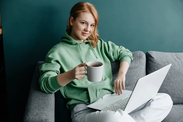 Ung rödhårig kaukasisk kvinna bär en grön luvtröja hemma shopping online med bärbar dator — Stockfoto