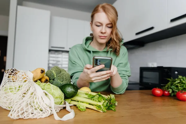 Nő használja okostelefon a modern konyhában, táska friss zöldséggel az asztalon. Összpontosítás az előtérben. Stock Kép