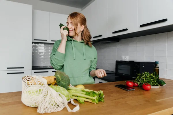 Mulher cheirando abacate fresco na cozinha moderna, saco com vegetais frescos na mesa. — Fotografia de Stock