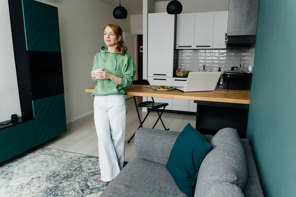 Young red-haired woman works at home and takes coffee break — Stock Photo, Image