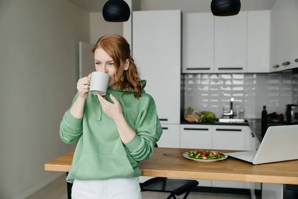 Genç kızıl saçlı kadın evde çalışıyor ve kahve molası veriyor. — Stok fotoğraf