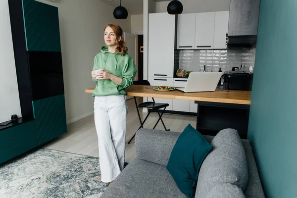 Young red-haired woman works at home and takes coffee break Royalty Free Stock Photos