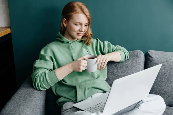 Ung rödhårig kaukasisk kvinna bär en grön luvtröja hemma shopping online med bärbar dator Royaltyfria Stockbilder
