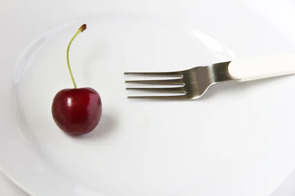 Uma cereja em uma chapa branca em um fundo branco e um garfo — Fotografia de Stock