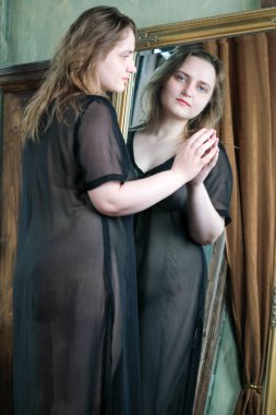A young pretty attractive brown-haired girl in a transparent black peignoir looks at her image in a large mirror