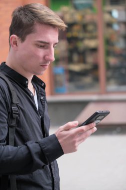 Portrait of a young handsome guy in a black shirt next to a brick wall with a smartphone autumn street photography