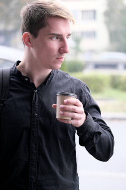 Portrait of a young handsome guy in a black shirt with a plastic glass of coffee autumn street photography