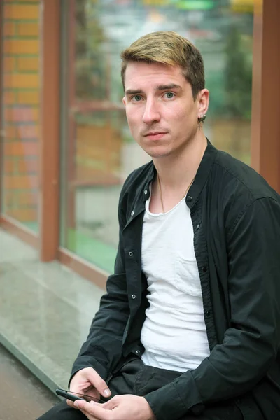 Portrait of a young handsome guy in a black shirt sits next to a shop window autumn street photography