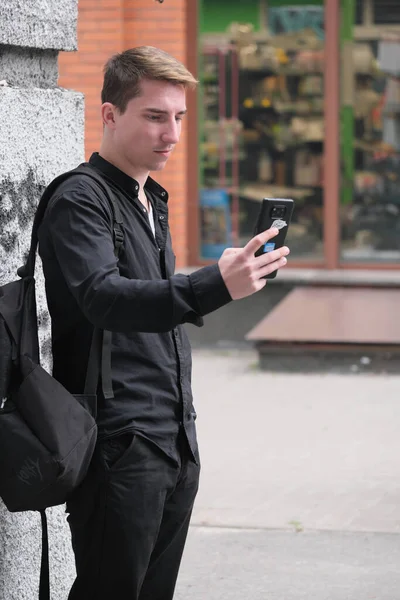 Portrait of a young handsome guy in a black shirt next to a brick wall with a smartphone autumn street photography
