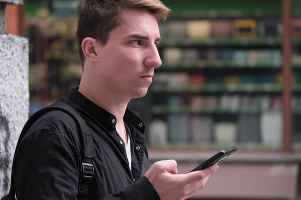 Portrait of a young handsome guy in a black shirt next to a brick wall with a smartphone autumn street photography