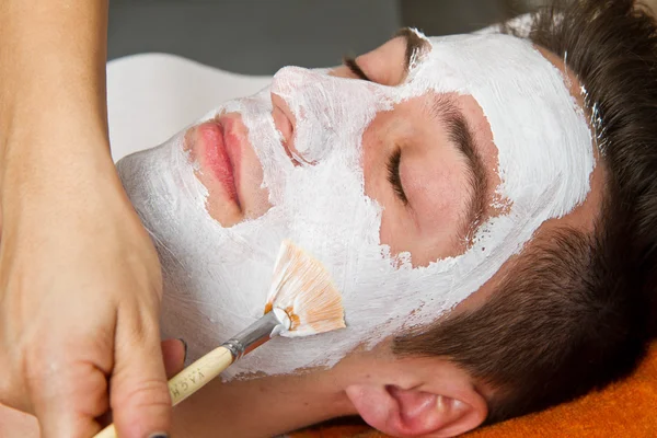 Θεραπευτής εφαρμόζει μια μάσκα προσώπου για ένα όμορφο νεαρό άνδρα σε ένα spa治疗师将口罩应用到一个美丽的年轻人，在一个水疗中心 — 图库照片