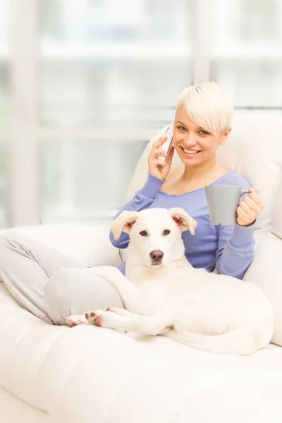 Mujer con perro en el sofá sosteniendo una taza y llamando —  Fotos de Stock
