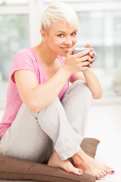 Mujer sonriente con taza sentada en casa mientras sonríe — Foto de Stock