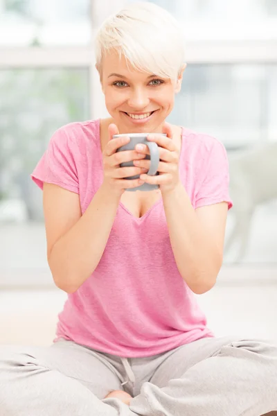 Mujer sonriente con taza sentada en casa mientras sonríe — Foto de Stock