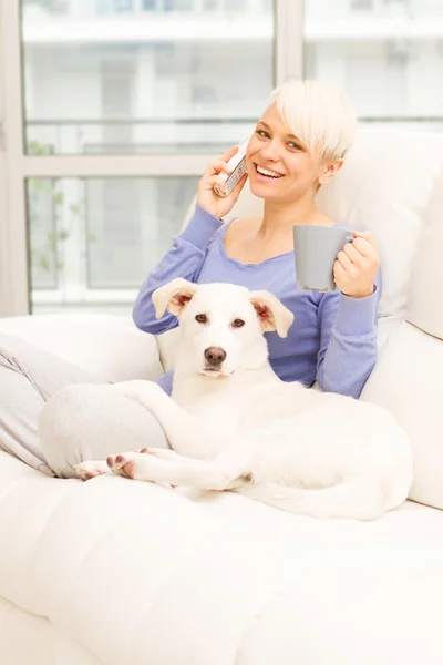 Frau mit Hund auf dem Sofa hält Becher in der Hand und telefoniert — Stockfoto