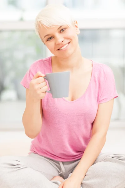Mujer sonriente con taza sentada en casa mientras sonríe — Foto de Stock