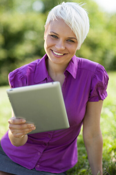 Smiling woman with tablet pc is sitting on the grass while smili