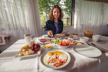 Bir kadın sabahları bir otel veya kafede doyurucu ve bol miktarda kıta kahvaltısı yer. Ön planda bir tabak kızarmış yumurta.