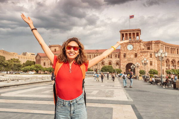 Happy student girl with a backpack raised her hands and rejoices wonderful view of the main tourist attraction of Yerevan - Republic Square