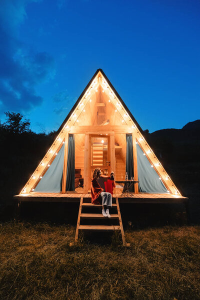 Woman drinking tea on the porch of a wooden lodge with lights of garlands in the evening. The concept of glamping and renting a chalet for weekend