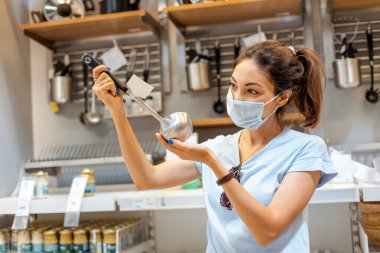Covid salgını sırasında koruyucu maskeli bir kadın alışveriş merkezinden bir kepçe ve diğer mutfak gereçlerini seçip alıyor.