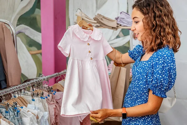 stock image Woman chooses a cute pink dress for her daughter in a boutique with a collection of children clothes. Shopping and motherhood concept