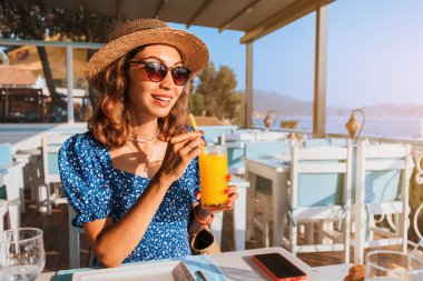 Şapkalı bir bayan gezgin, deniz kıyısına bakan bir barın çatısında portakal suyuyla kokteyl içiyor..