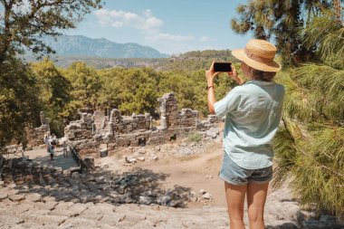 Türkiye 'nin antik Phaselis kentindeki Roma amfi tiyatrosunun kalıntılarının akıllı telefonuyla fotoğraf çeken turist kız. Gezmek ve gezmek