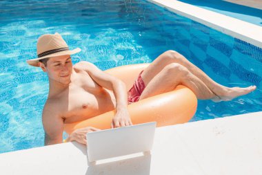 Relaxed man floating with inflatable circle in swimming pool and surfing the internet websites at laptop or notebook pc. Leisure weekend and remote freelance work
