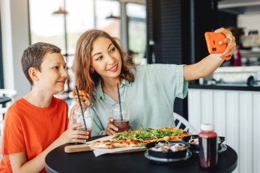 Anne ve oğlu bir kafede pizza yerken ve sosyal ağlar için akıllı telefondan selfie çekerken iyi vakit geçiriyorlar.