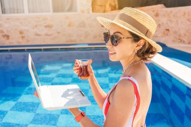 Laptop bilgisayarı ve tatil köyündeki bir havuzun arka planında bir bardak Türk çayı. Uzak serbest çalışma ve hafta sonu konsepti