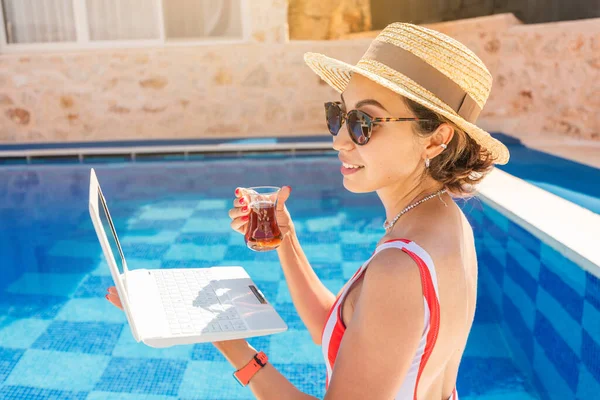 Laptop bilgisayarı ve tatil köyündeki bir havuzun arka planında bir bardak Türk çayı. Uzak serbest çalışma ve hafta sonu konsepti