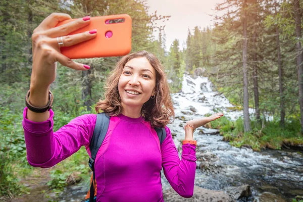 一位女性旅行博客作者在她的手机上为社交网络拍了个自拍 背景是一场精彩的瀑布 — 图库照片